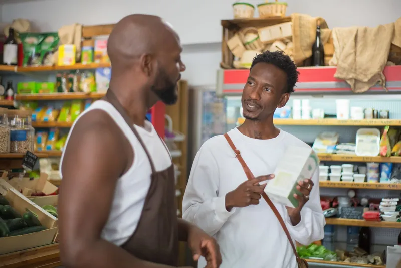 A shopkeeper in an apron talking a customer through a product in a small grocery store