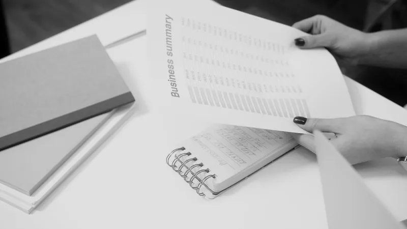Hands turning the pages of a printed business summary on a white desk